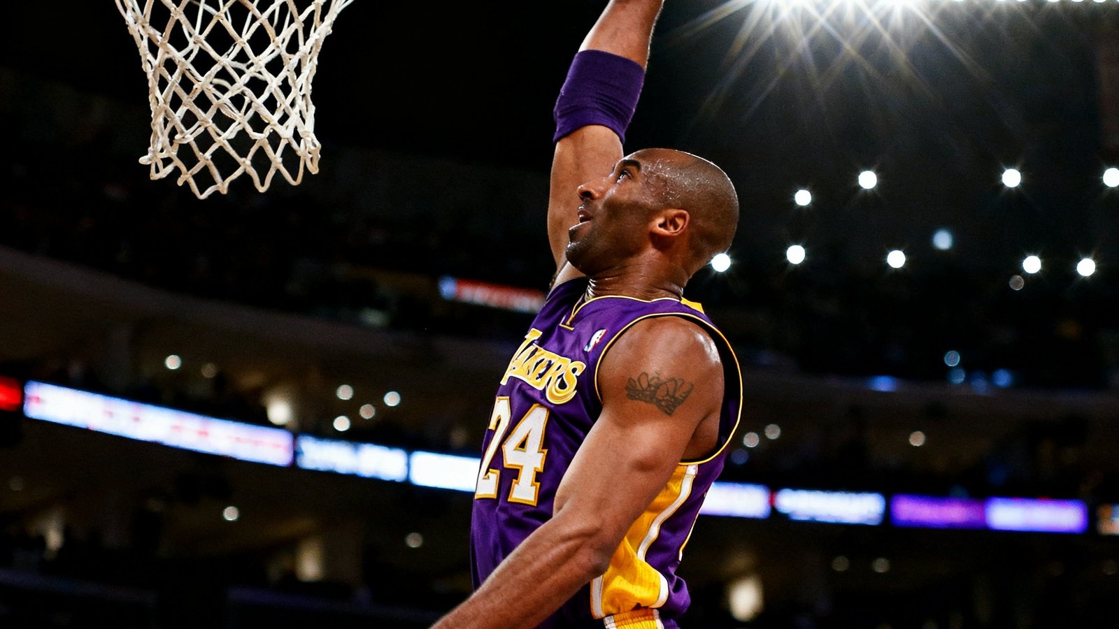 NBA赛场上身穿紫金球衣的球员飞身扣篮，手臂高举篮球即将砸入篮筐，背景中对手球员仰头目送，球馆内闪光灯密集亮起